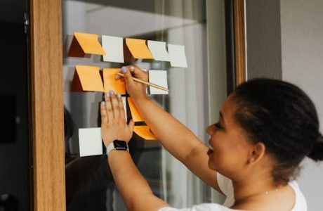 A woman writes notes on a window using colorful sticky notes, emphasizing planning and creativity.